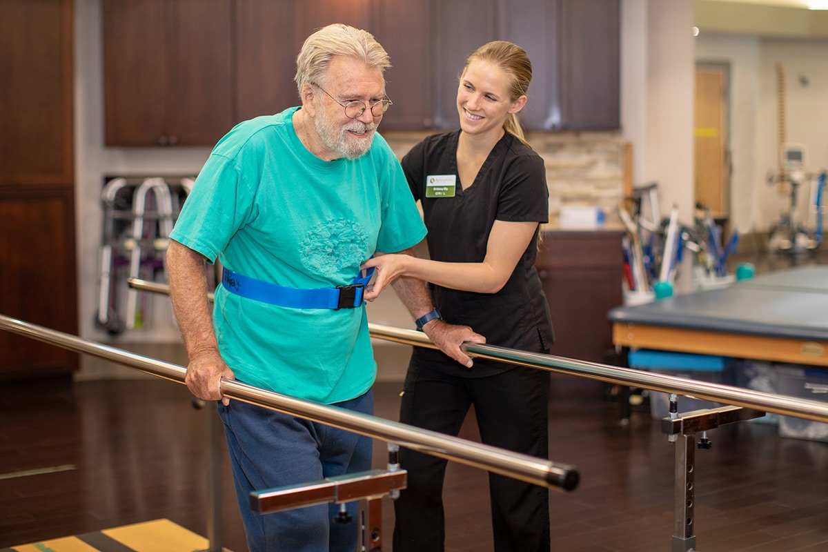 Therapist in black scrubs and blonde ponytail helping man in blue shirt
