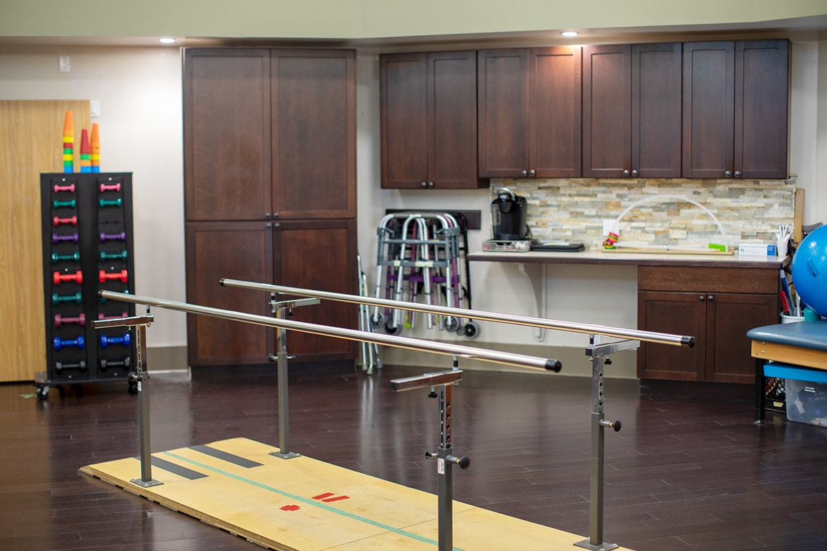 Rehab gym with dark wood floors, parallel bars and other rehab equipment.