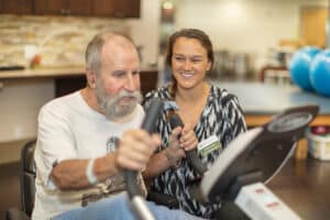 Therapist helping elderly man with beard use rehab bike in the rehab gym.