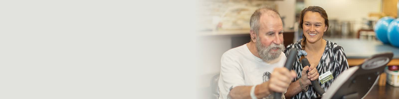 Therapist helping elderly man with beard use rehab bike in the rehab gym at Redwood Cove Healthcare